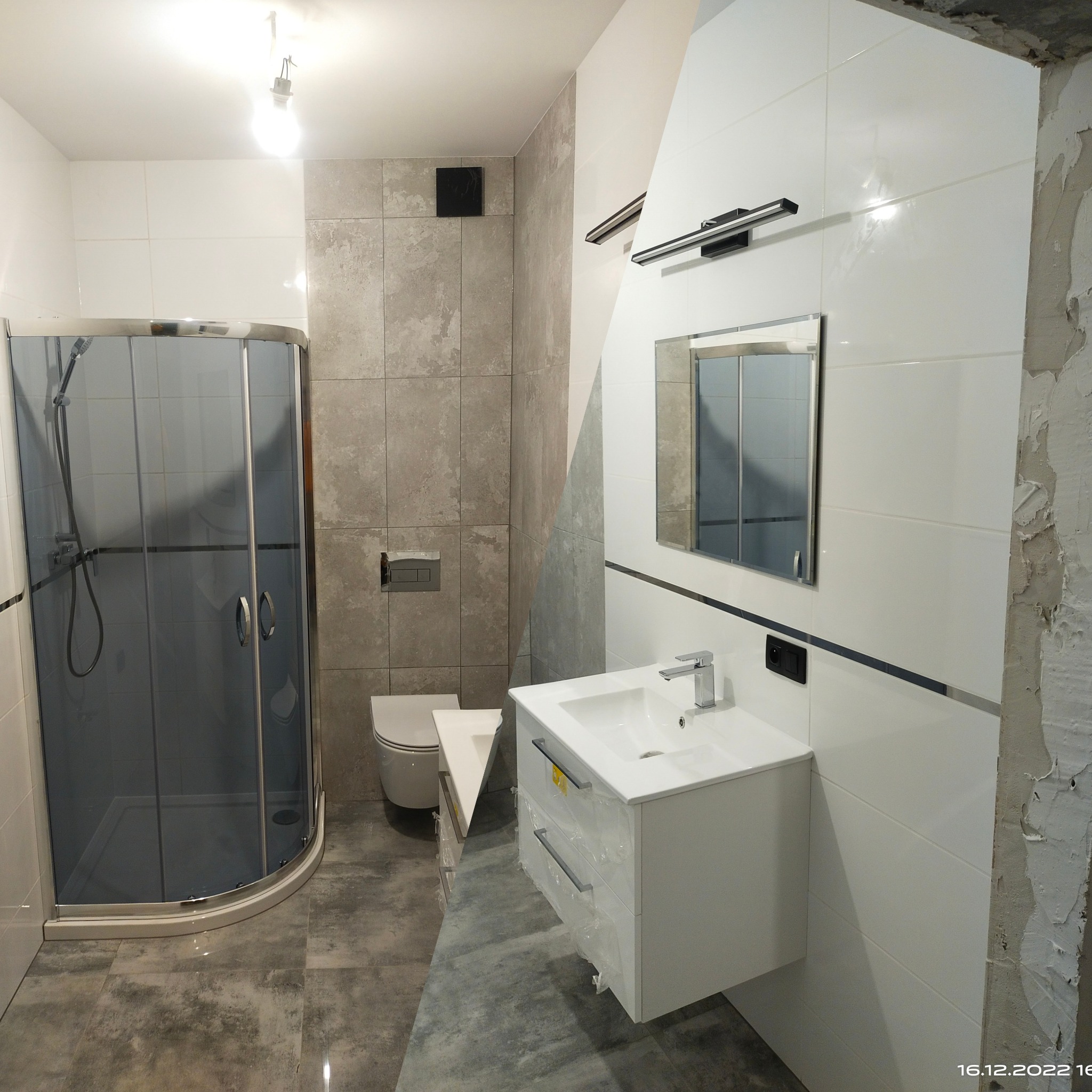 Nowoczesna łazienka w trakcie wykańczania: kabina prysznicowa z szarymi szybami, umywalka z szafką w białym połysku, szare płytki imitujące kamień na ścianie, białe płytki z czarnym paskiem...