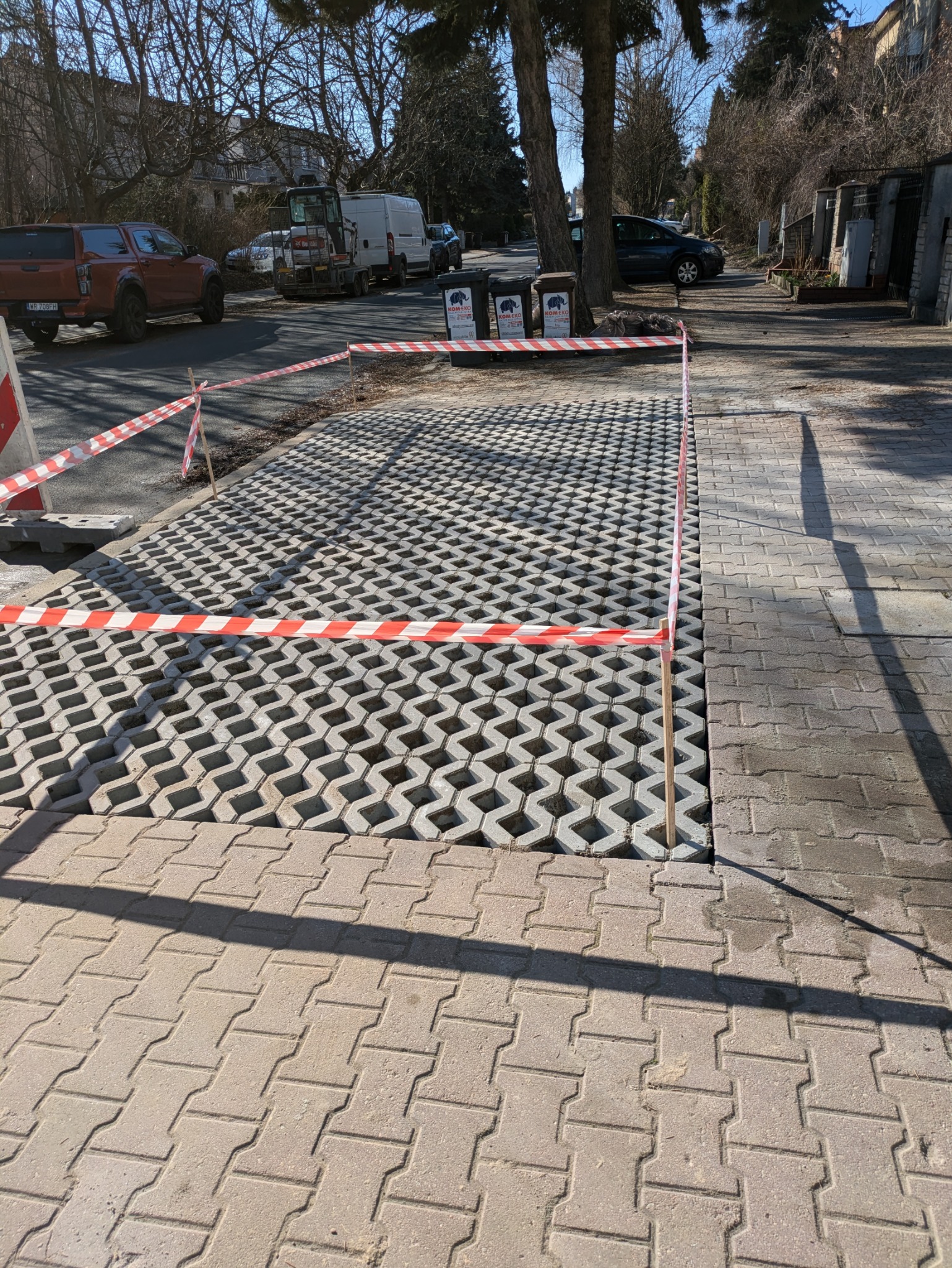 Układanie przepuszczalnej kostki brukowej z otworami, oddzielonej taśmą ostrzegawczą od istniejącego chodnika z klasycznej kostki, widoczne samochody i sprzęt budowlany w tle.