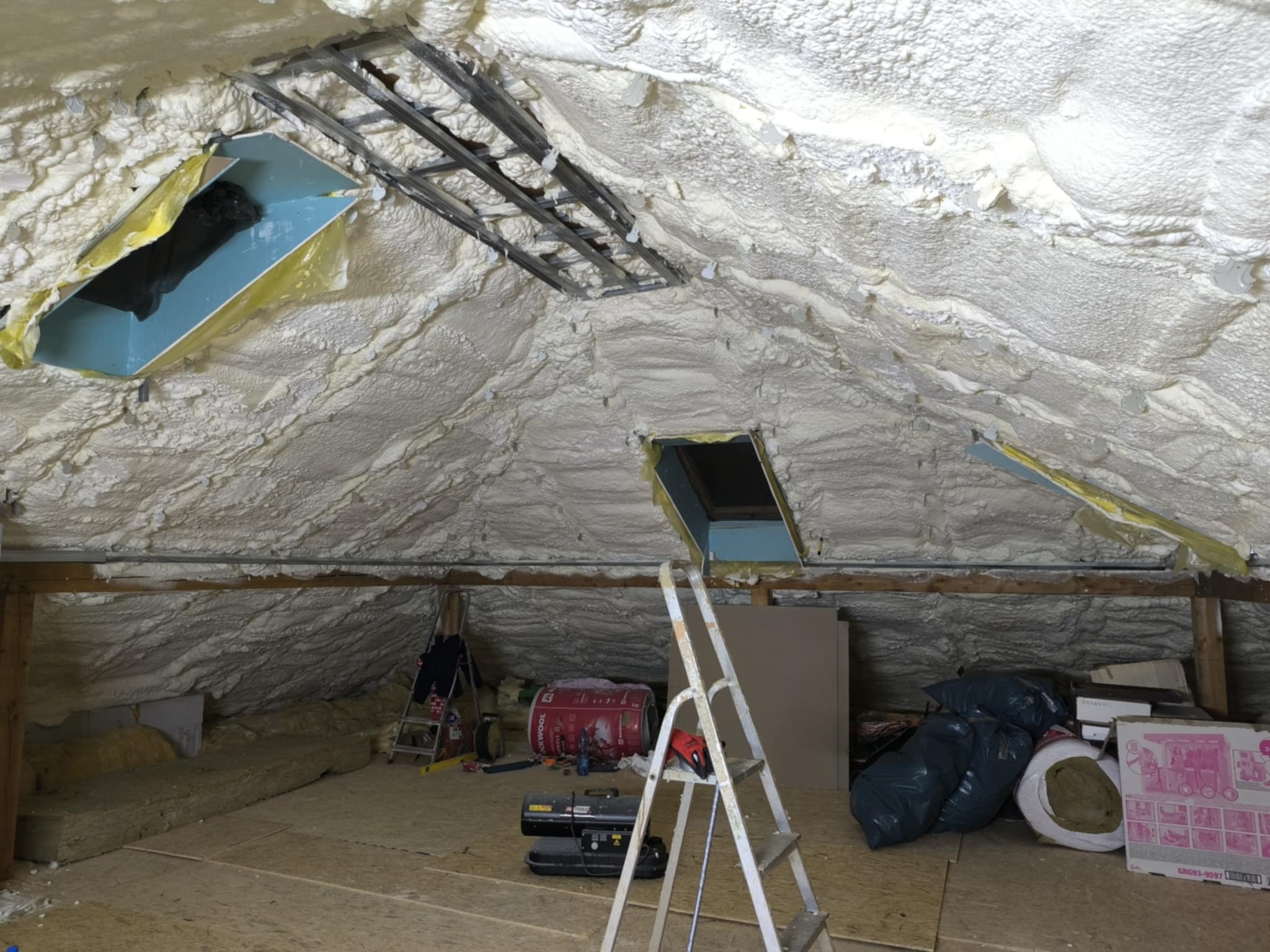 Poddasze po ociepleniu pianką PUR, widoczne okna dachowe i konstrukcja metalowa. Na podłodze narzędzia i materiały budowlane, drabina. Ocieplenie wykonane natryskowo.