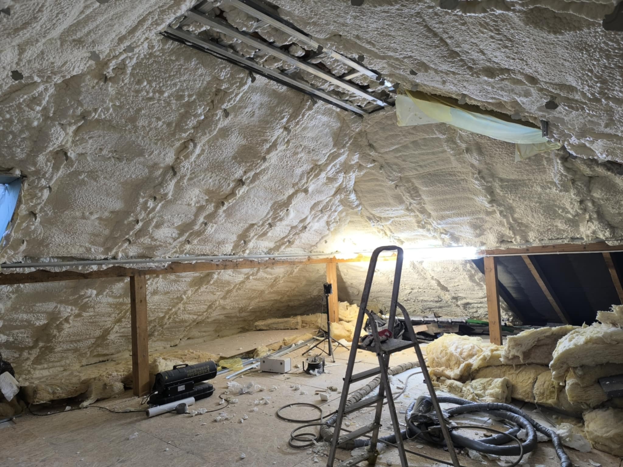 Poddasze po ociepleniu pianką PUR: biała, nierówna powierzchnia pokrywa skośny dach i ściany. Widoczne elementy konstrukcyjne i narzędzia pracy.