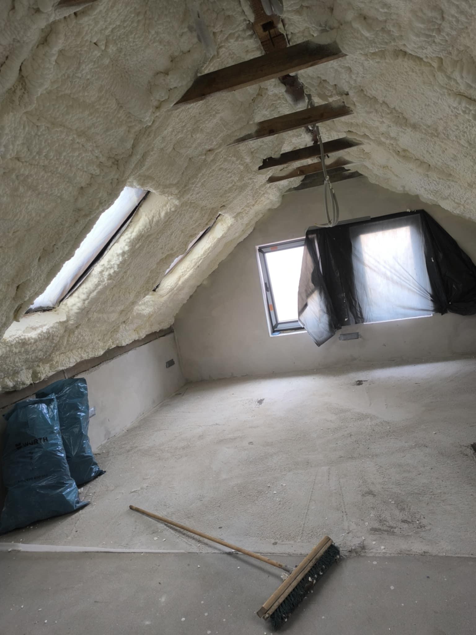 Poddasze po ociepleniu pianką PUR, widoczne worki z odpadami i szczotka. Pianka pokrywa skosy i okna dachowe. Jeden z okien zasłonięty folią.