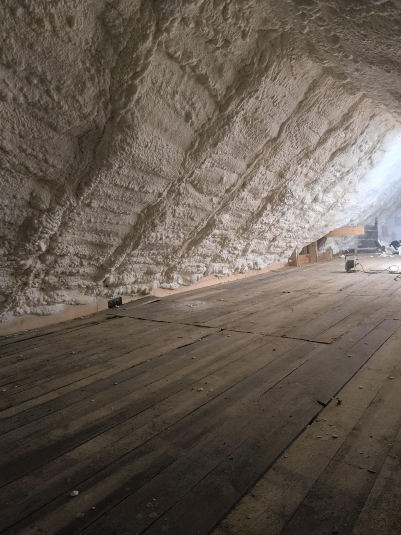 Ocieplanie poddasza pianką PUR w Prudniku. Widoczne drewniane deski podłogowe i biała pianka na skośnej ścianie poddasza. Charakterystyczna faktura piany.