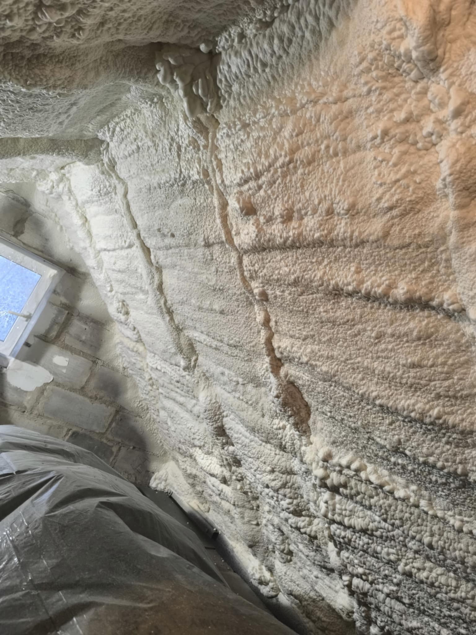 Poddasze ocieplone pianką PUR, widoczne nierówności i struktura piany. Fragment okna i zabezpieczona folią podłoga. Ocieplenie natryskowe.