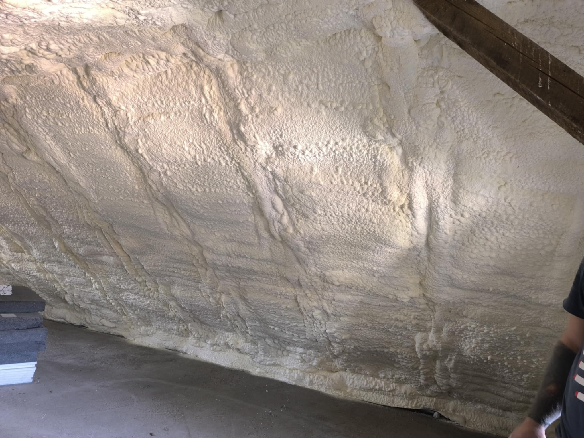 Ocieplenie poddasza pianką PUR, widoczne nierówności struktury piany. Fragment drewnianej belki stropowej w prawym górnym rogu. Szara podłoga i ułożone płyty izolacyjne.