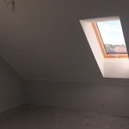 Poddasze ze skośnym sufitem i oknem dachowym z widokiem na zabudowę jednorodzinną, ściany po świeżym gruntowaniu przed malowaniem, surowy beton na podłodze.