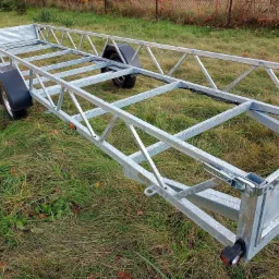 Szkielet metalowej przyczepki samochodowej z ocynkowaną ramą kratownicową, widoczne koła z błotnikami i zapięcia burty, tło stanowi trawa.