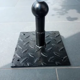 Metalowy element konstrukcyjny z kulistym zakończeniem, przyspawany do kwadratowej podstawy z wzorem łezkowym, całość pomalowana na czarno i umieszczona na szarej, płytkowej powierzchni.