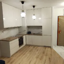 Nowoczesna kuchnia z białymi, lakierowanymi frontami, drewnianym blatem i marmurowym backsplashiem; wiszące lampy z ryflowanego szkła, widok na wejście z zabezpieczonymi drzwiami.