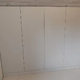 Biała szafa wnękowa z czterema frontami, wbudowana pod skosem drewnianego sufitu, z widocznymi gniazdkami elektrycznymi na ścianie i podłodze.