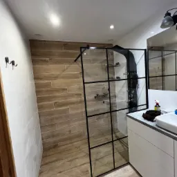 Nowoczesna kabina prysznicowa typu walk-in z czarnymi profilami i deszczownicą, wykończona płytkami imitującymi drewno, z widoczną umywalką i szafką w tle.