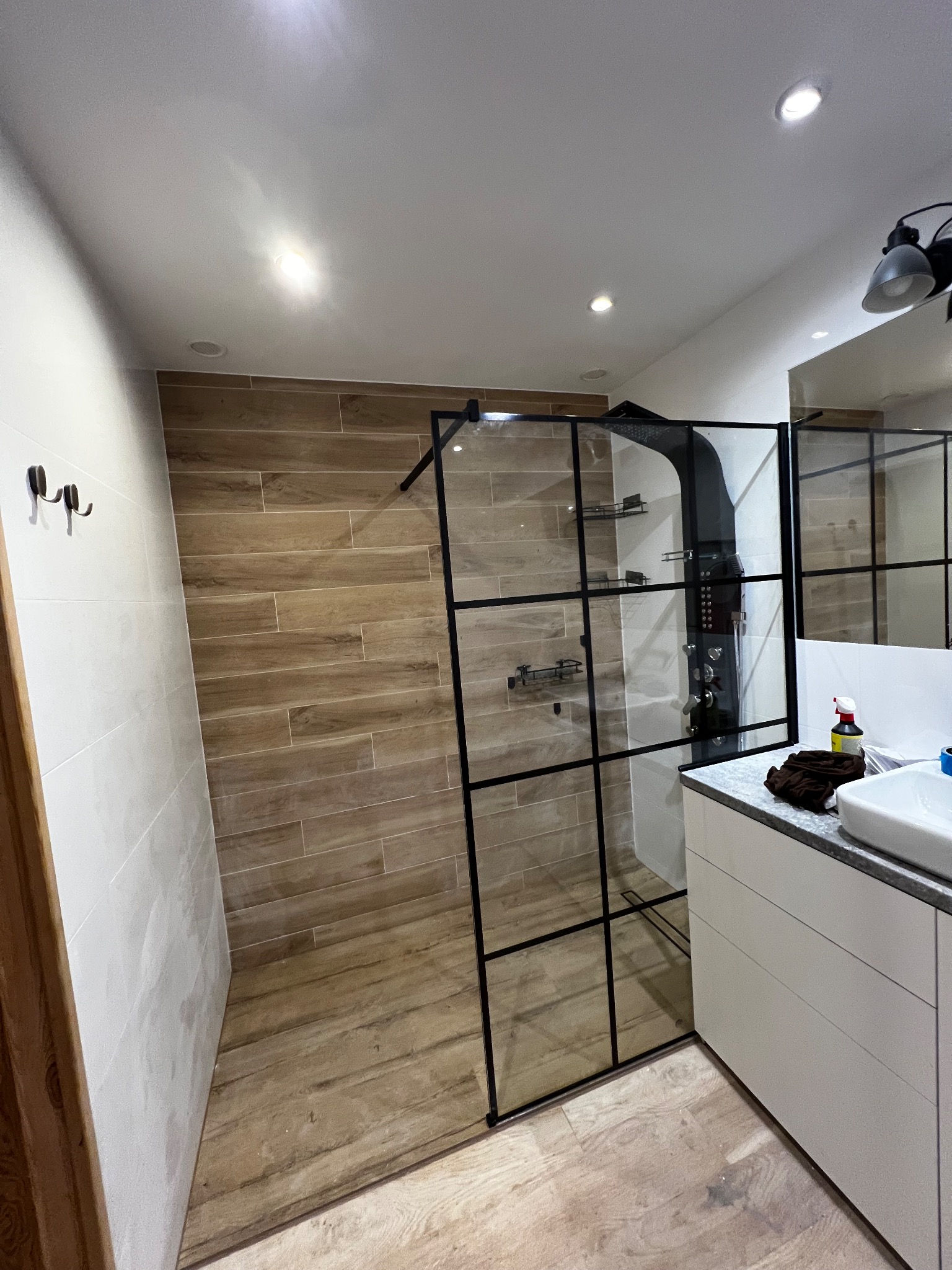 Nowoczesna kabina prysznicowa typu walk-in z czarnymi profilami i deszczownicą, wykończona płytkami imitującymi drewno, z widoczną umywalką i szafką w tle.