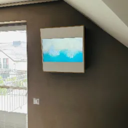 Biała klimatyzacja kasetonowa LG Artcool Gallery z wyświetlanym obrazem plaży, zamontowana na brązowej ścianie w pokoju z oknem i roletami.