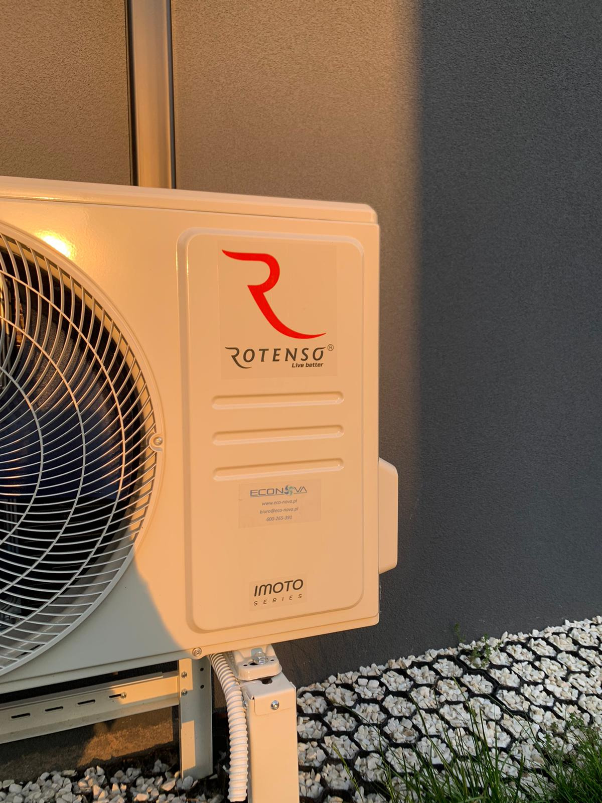 Zewnętrzna jednostka klimatyzacji Rotenso Imoto Series zamontowana przy szarej ścianie budynku, oświetlona ciepłym światłem zachodzącego słońca, widoczna kratka z kamieniami u podstawy.