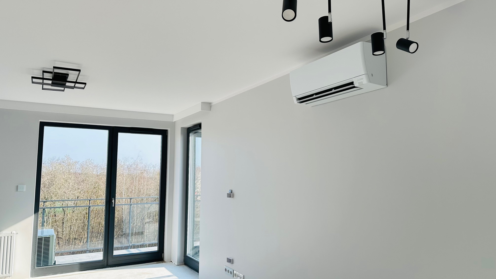 Wnętrze jasnego pokoju z zamontowanym na ścianie, białym klimatyzatorem Mitsubishi, widoczne czarne lampy sufitowe i okno balkonowe z widokiem na drzewa.