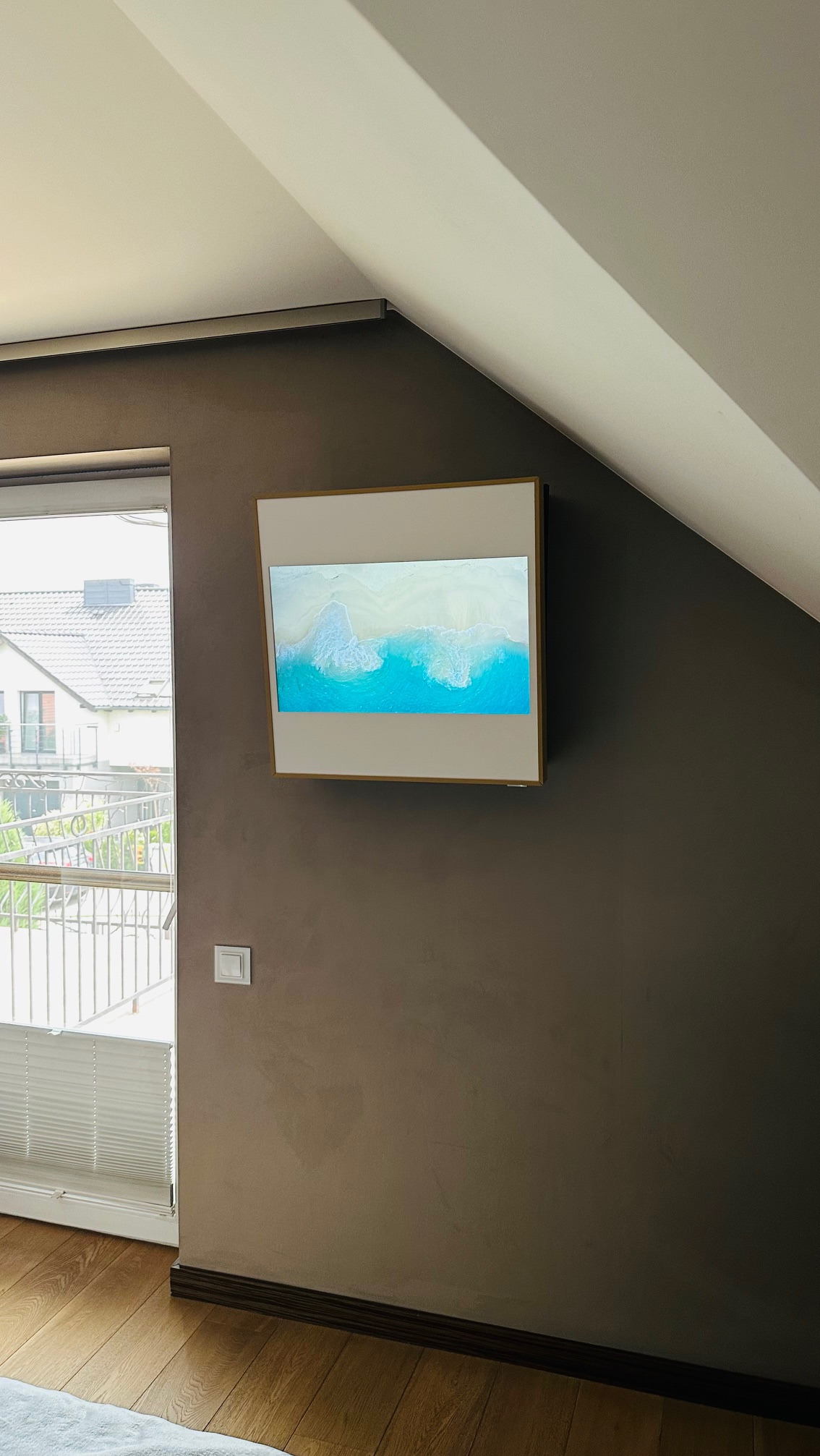 Biała klimatyzacja kasetonowa LG Artcool Gallery z wyświetlanym obrazem plaży, zamontowana na brązowej ścianie w pokoju z oknem i roletami.