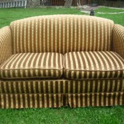 Dwuosobowa sofa w brązowo-beżowe pionowe pasy, z dekoracyjnymi guzikami na bokach i falbaną u dołu, ustawiona na zielonej trawie.