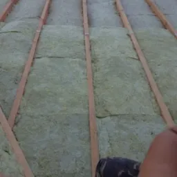 Izolacja termiczna dachu z wełny mineralnej pomiędzy drewnianymi krokwiami, widoczne fragmenty ubrania osoby wykonującej prace.