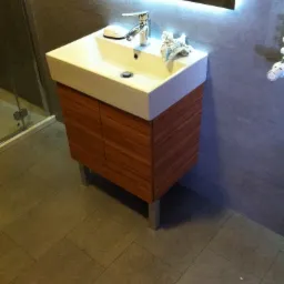 Nowoczesna łazienka z białą umywalką nablatową na szafce z drewnianymi frontami, metalowe nóżki, szare płytki na ścianach i podłodze, oświetlenie LED nad lustrem.