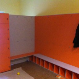 MR Meble Magdalena Okońska - Pomarańczowo-biała szatnia z szafkami i wieszakami w przedszkolu lub szkole, z żółtą ścianą i podłogą w odcieniach brązu.