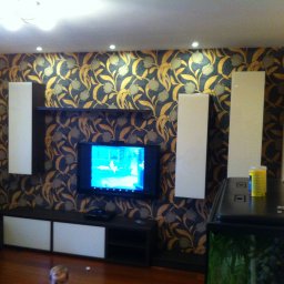 MR Meble Magdalena Okońska - Telewizor na ścianie z tapetą w kwiaty, obok pionowe białe panele i szafki RTV w salonie, na podłodze widoczna głowa dziecka.