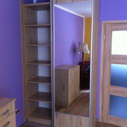 MR Meble Magdalena Okońska - Jasna szafa wnękowa z przesuwnymi drzwiami z lustrem, widoczne półki wewnątrz, na podłodze panele, ściany fioletowe, drzwi drewniane z szybą.