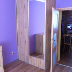 MR Meble Magdalena Okońska - Szafa wnękowa z przesuwnymi drzwiami o drewnianym wzorze, wnęka z półką, drewniana podłoga i otwarte drzwi do pokoju z widocznym telewizorem.