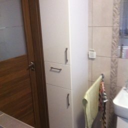 MR Meble Magdalena Okońska - Biała, wysoka szafa łazienkowa z trzema segmentami i metalowymi uchwytami, stojąca obok drewnianych drzwi w łazience wyłożonej beżowymi płytkami.