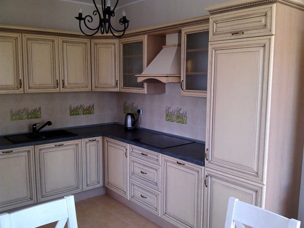 Klasyczna kuchnia z kremowymi frontami szafek, ciemnym blatem i dekoracyjnymi płytkami z motywem lawendy, oświetlona czarnym żyrandolem.