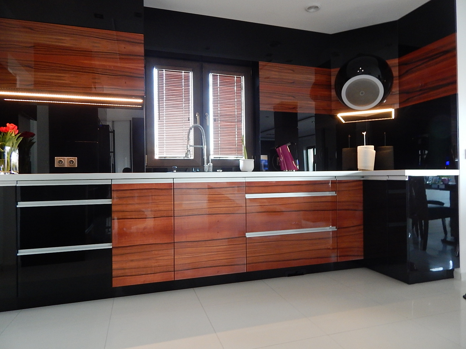 Nowoczesna kuchnia z frontami z forniru o ciepłym odcieniu drewna, czarnymi błyszczącymi elementami i białym blatem, oświetlona ledowym paskiem pod szafkami wiszącymi, z widocznym zlewem pod oknem...