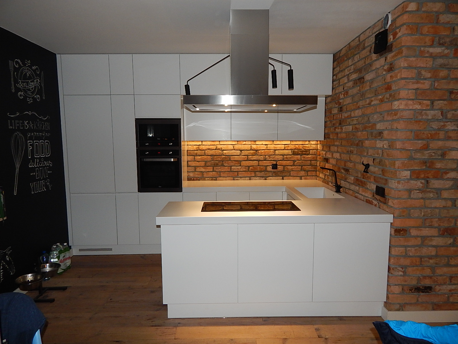 Nowoczesna kuchnia z białymi frontami i ceglaną ścianą, oświetlona listwą LED pod szafkami wiszącymi, z czarną tablicą kredową z rysunkami po lewej stronie.