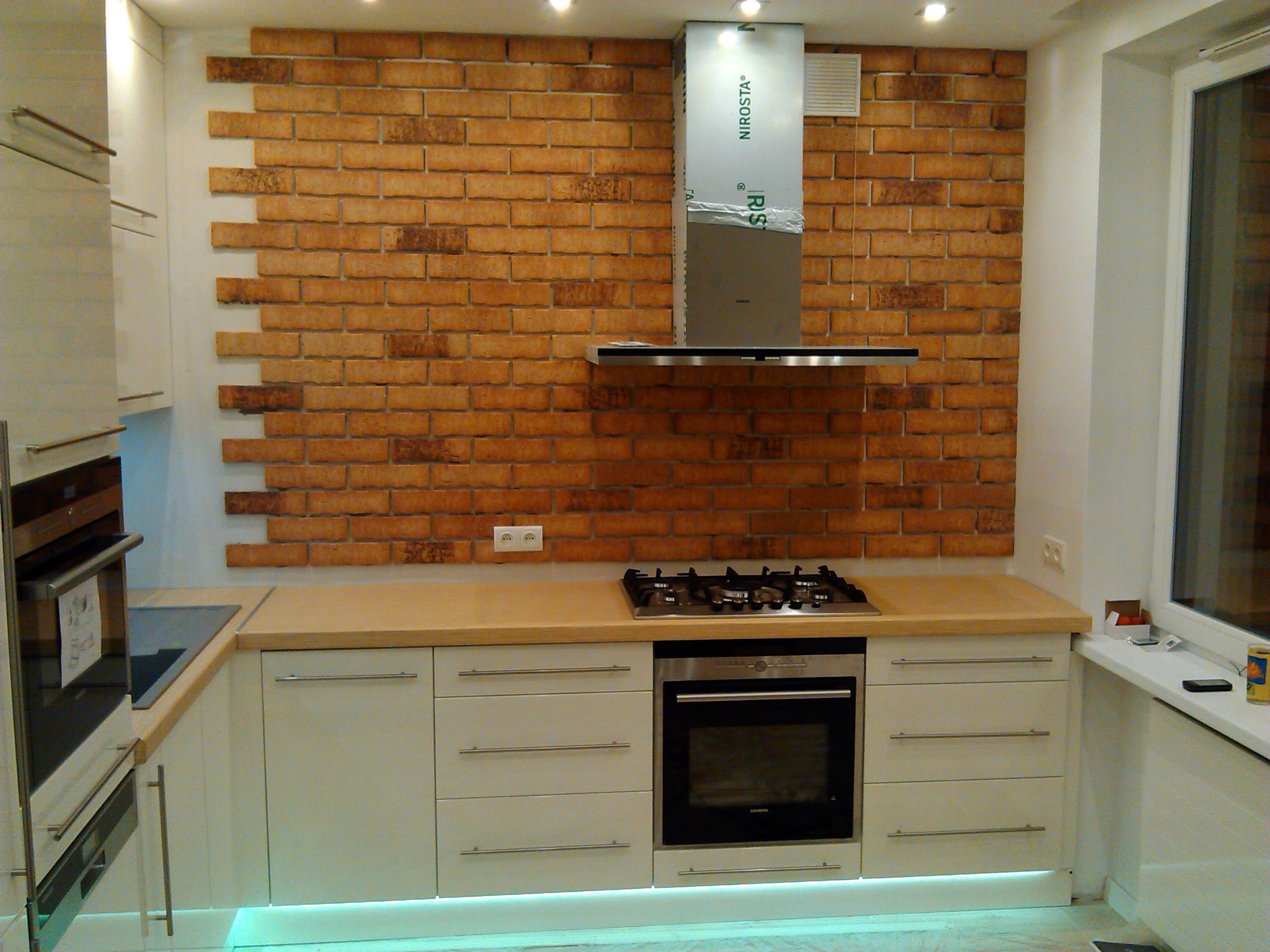 Nowoczesna kuchnia z białymi frontami szafek, blatem w odcieniach beżu i ścianą wyłożoną cegłą, oświetlenie LED pod szafkami, płyta gazowa i piekarnik w zabudowie.