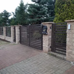 Nowoczesne poziome przęsła w kolorze antracytowym, osadzone na słupkach z imitacji piaskowca, brama wjazdowa z domofonem i skrzynką na listy wkomponowana w ogrodzenie posesji z brukowanym podjazdem.