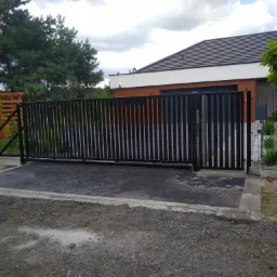 Nowoczesna, przesuwna brama wjazdowa z pionowymi, czarnymi profilami na posesji z kostką brukową i trawnikiem, widok z zewnątrz.