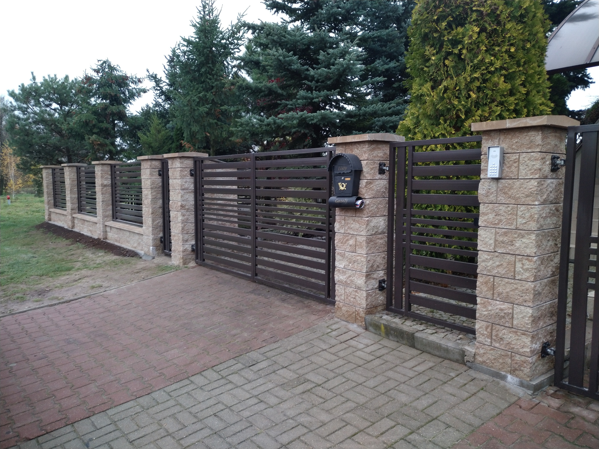 Nowoczesne poziome przęsła w kolorze antracytowym, osadzone na słupkach z imitacji piaskowca, brama wjazdowa z domofonem i skrzynką na listy wkomponowana w ogrodzenie posesji z brukowanym podjazdem.
