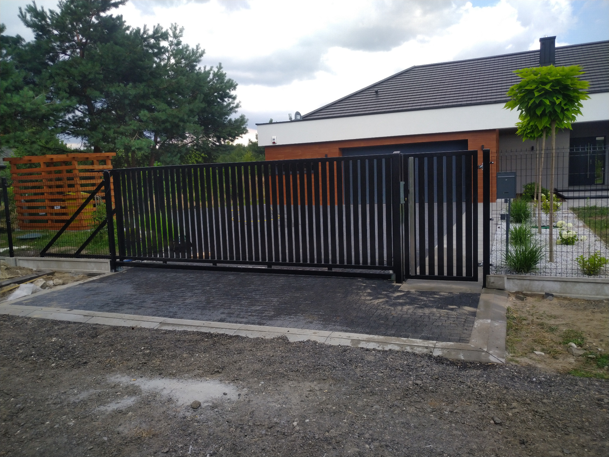 Nowoczesna, przesuwna brama wjazdowa z pionowymi, czarnymi profilami na posesji z kostką brukową i trawnikiem, widok z zewnątrz.