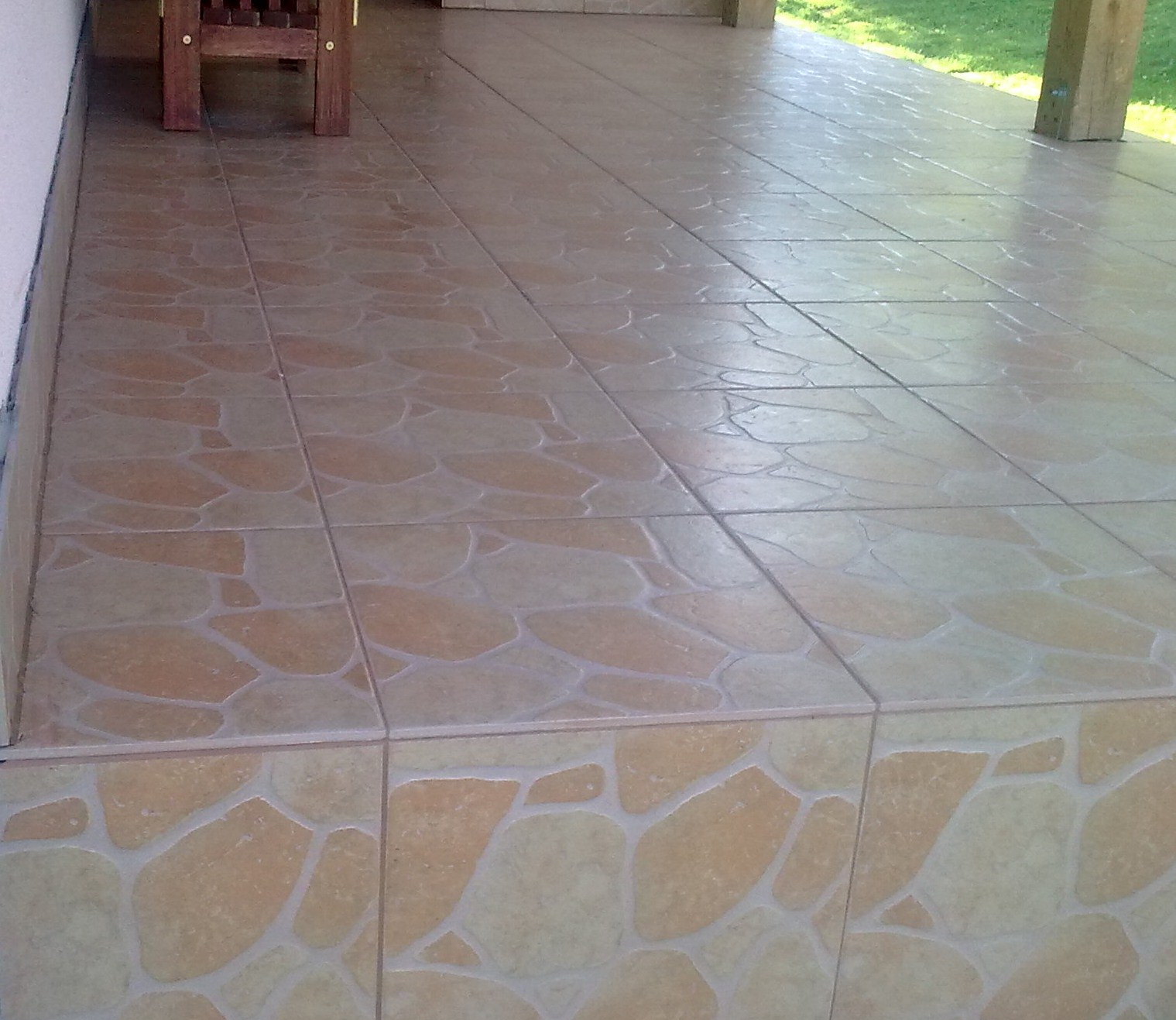 Wyłożona płytkami podłoga na zewnątrz budynku, z widocznym fragmentem drewnianego mebla i zieleni w tle.