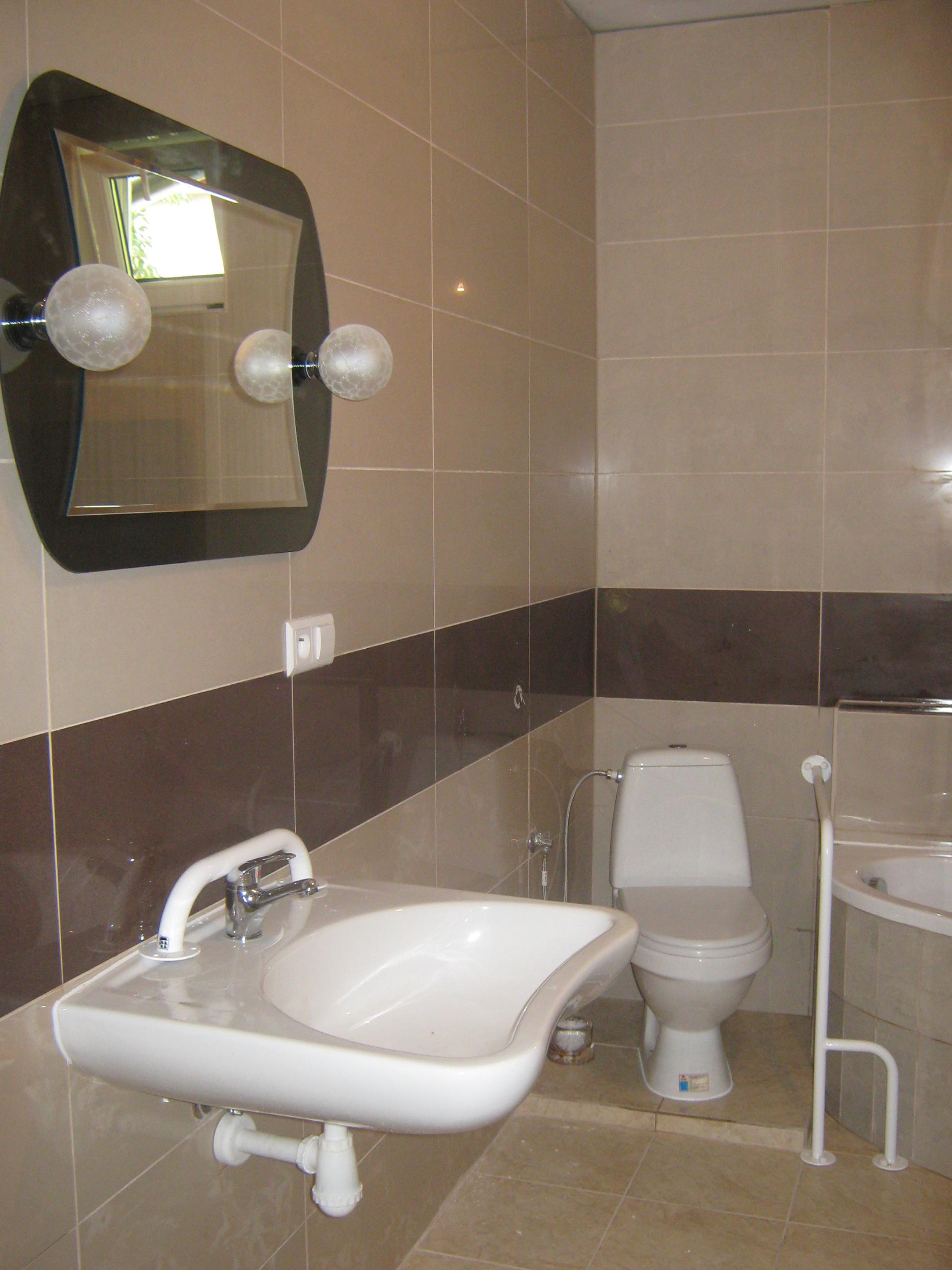 Umywalka z chromowaną baterią i uchwytem dla niepełnosprawnych w wykafelkowanej łazience z toaletą i narożną wanną; na ścianie lustro z kulistymi lampami.
