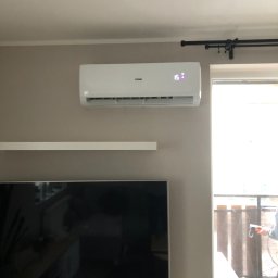 Props Klima Mateusz Kawczyński - Biały klimatyzator Haier zamontowany na ścianie w salonie, wyświetlający temperaturę 15 stopni, obok okna balkonowego z widocznym fragmentem balkonu.