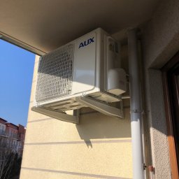Props Klima Mateusz Kawczyński - Biały klimatyzator AUX zamontowany na zewnątrz budynku mieszkalnego, widoczne rury odprowadzające, budynek w tle.