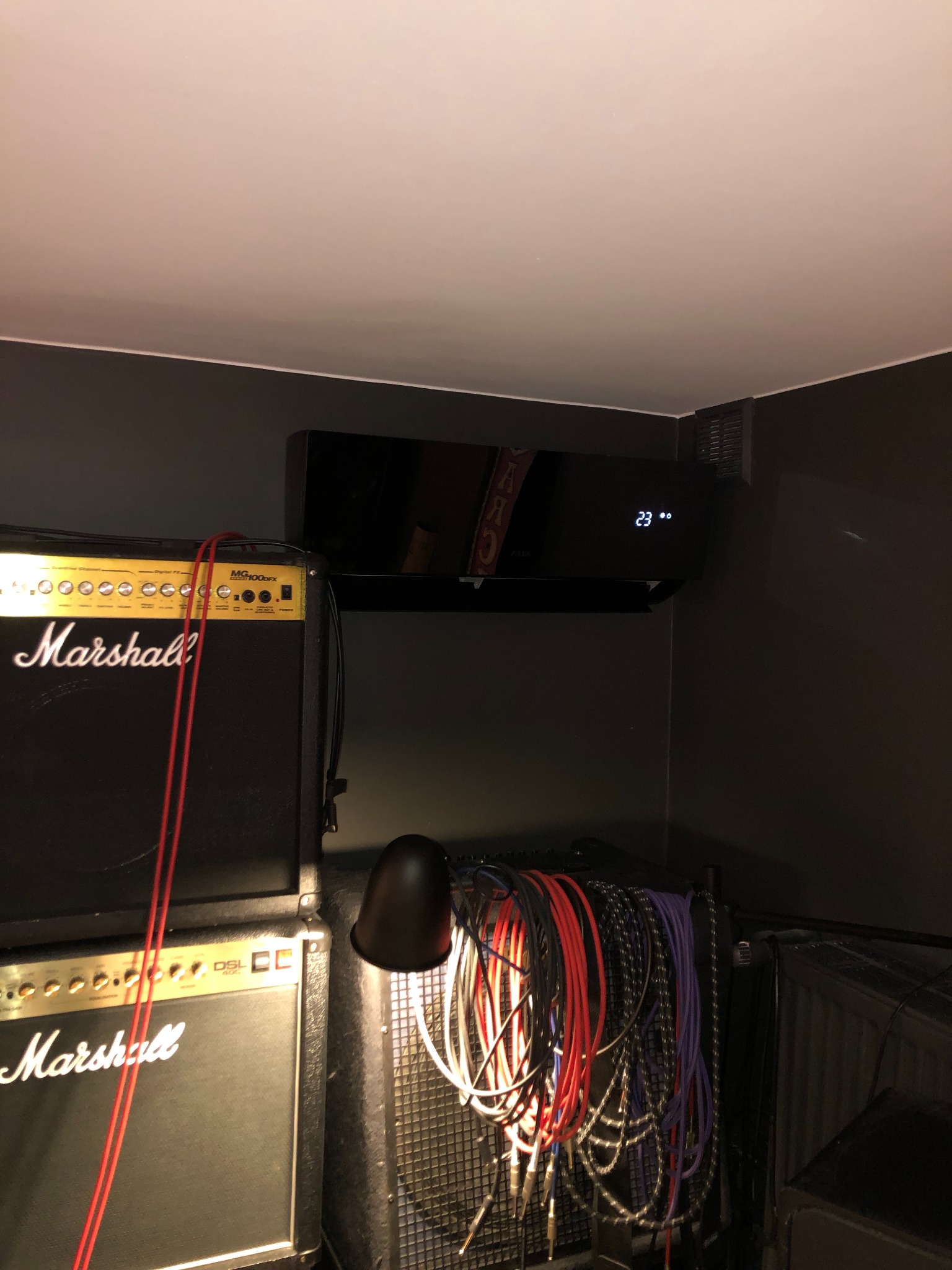 Czarny klimatyzator ścienny zamontowany w pokoju muzycznym, widoczne wzmacniacze gitarowe Marshall i plątanina kabli.