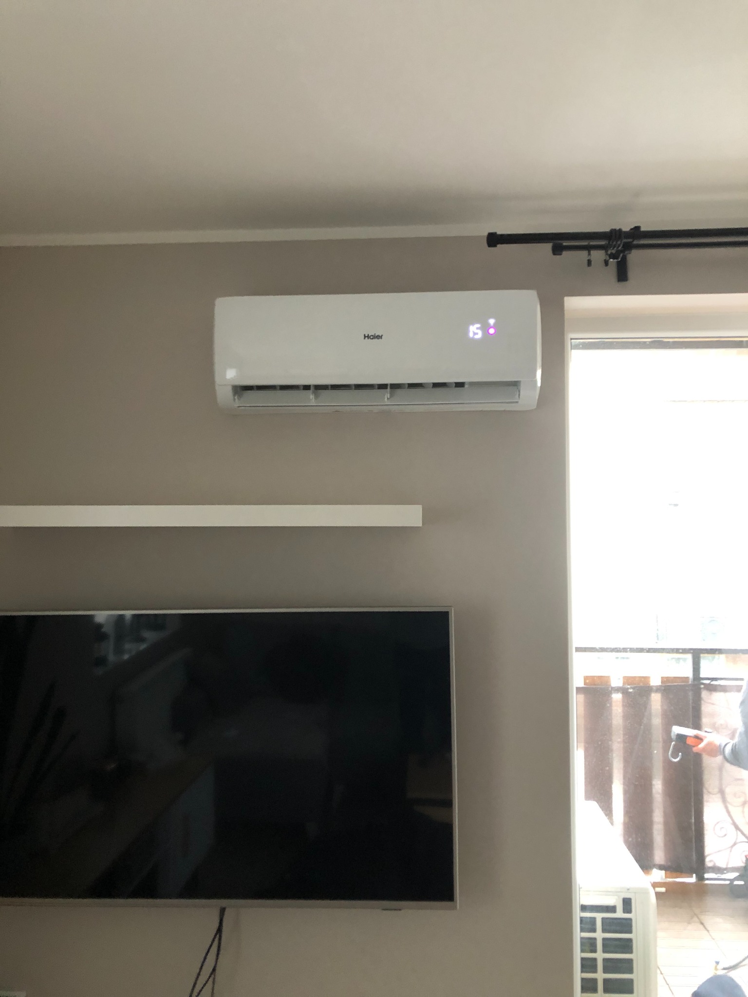 Biały klimatyzator Haier zamontowany na ścianie w salonie, wyświetlający temperaturę 15 stopni, obok okna balkonowego z widocznym fragmentem balkonu.