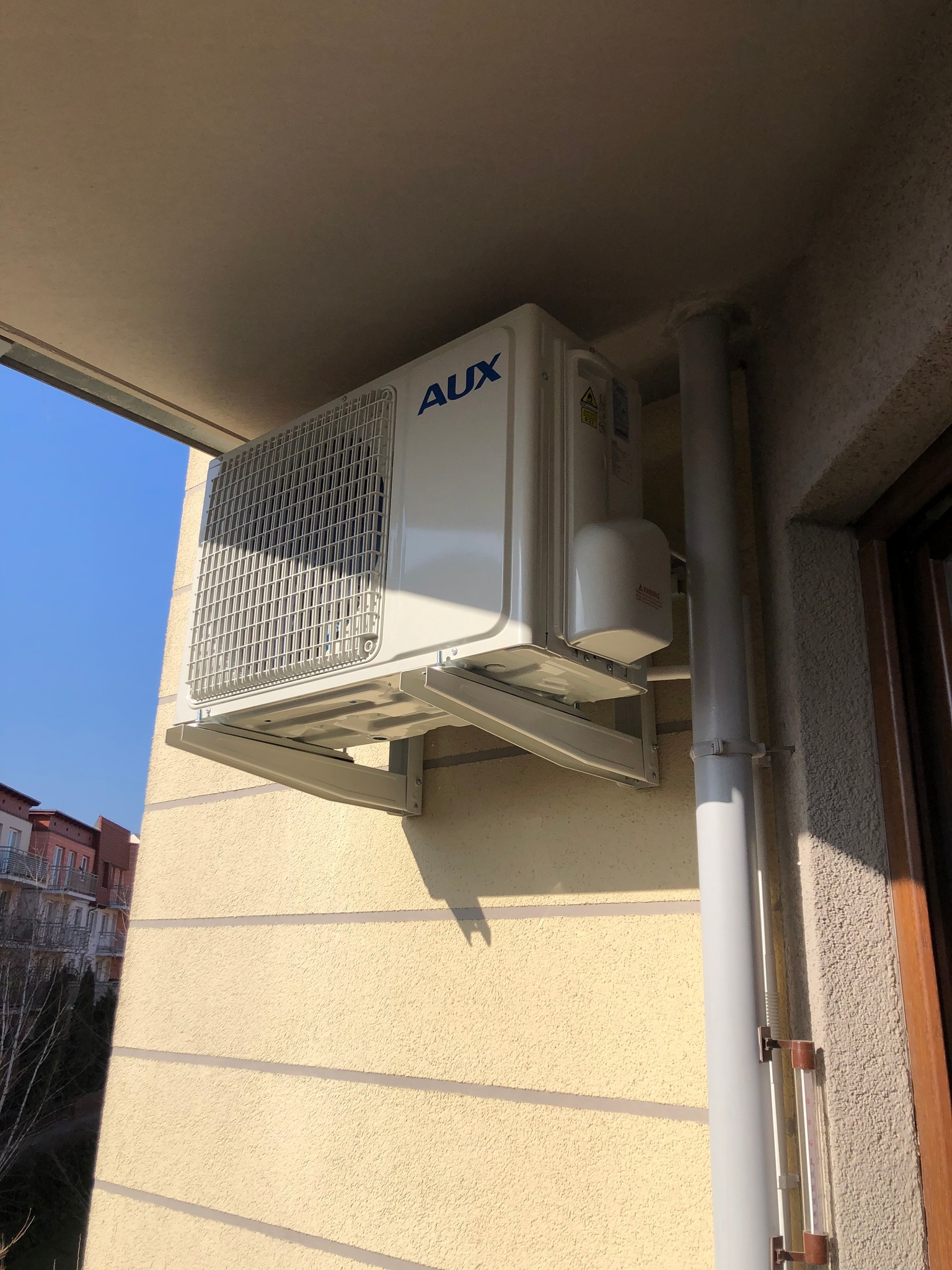 Biały klimatyzator AUX zamontowany na zewnątrz budynku mieszkalnego, widoczne rury odprowadzające, budynek w tle.