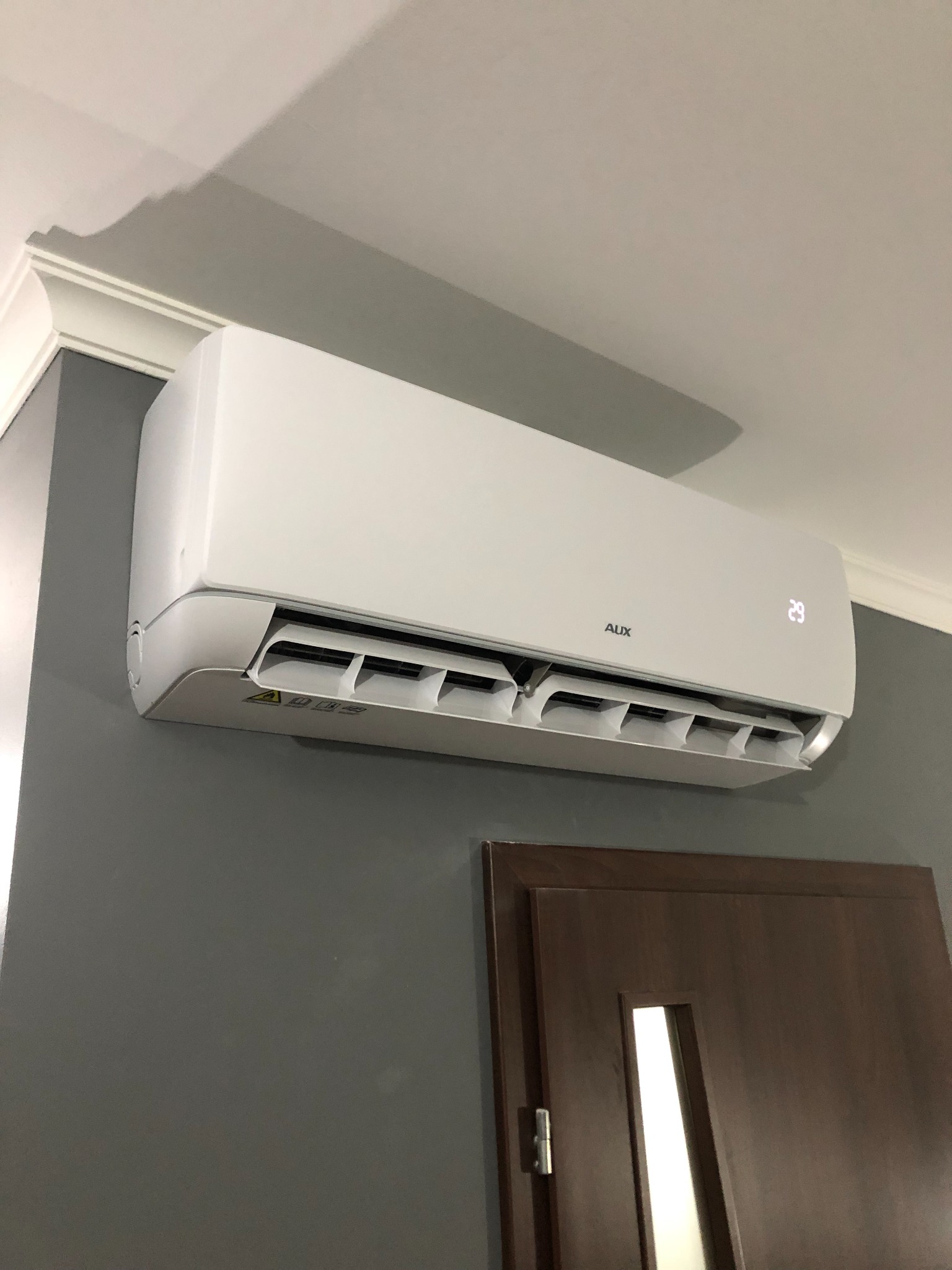 Biała klimatyzacja AUX zamontowana na szarej ścianie w pomieszczeniu z brązowymi drzwiami, wyświetlająca temperaturę 29 stopni Celsjusza.