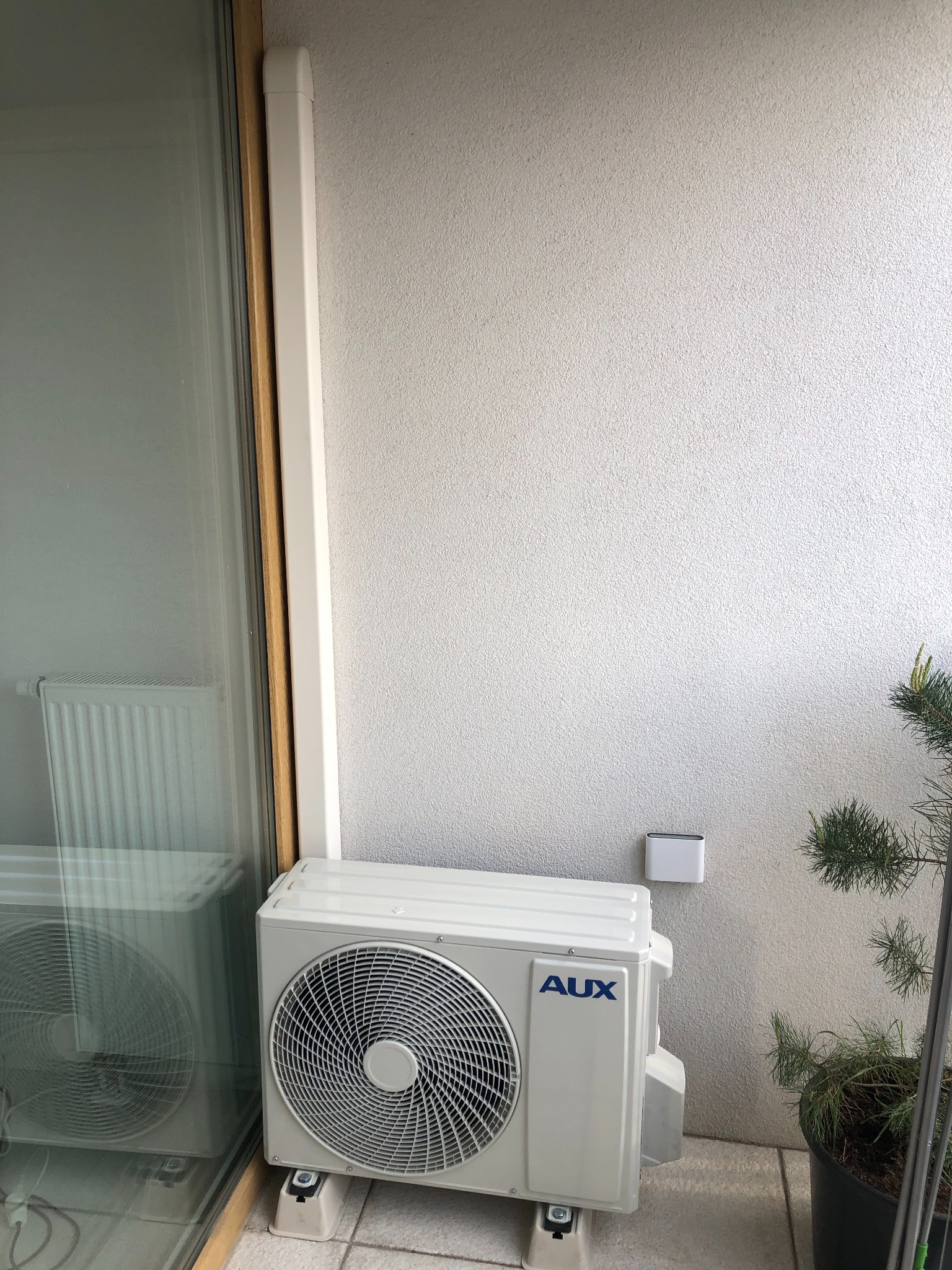Biała jednostka zewnętrzna klimatyzacji AUX zamontowana na balkonie, widoczna instalacja odprowadzająca skropliny wzdłuż okna balkonowego.