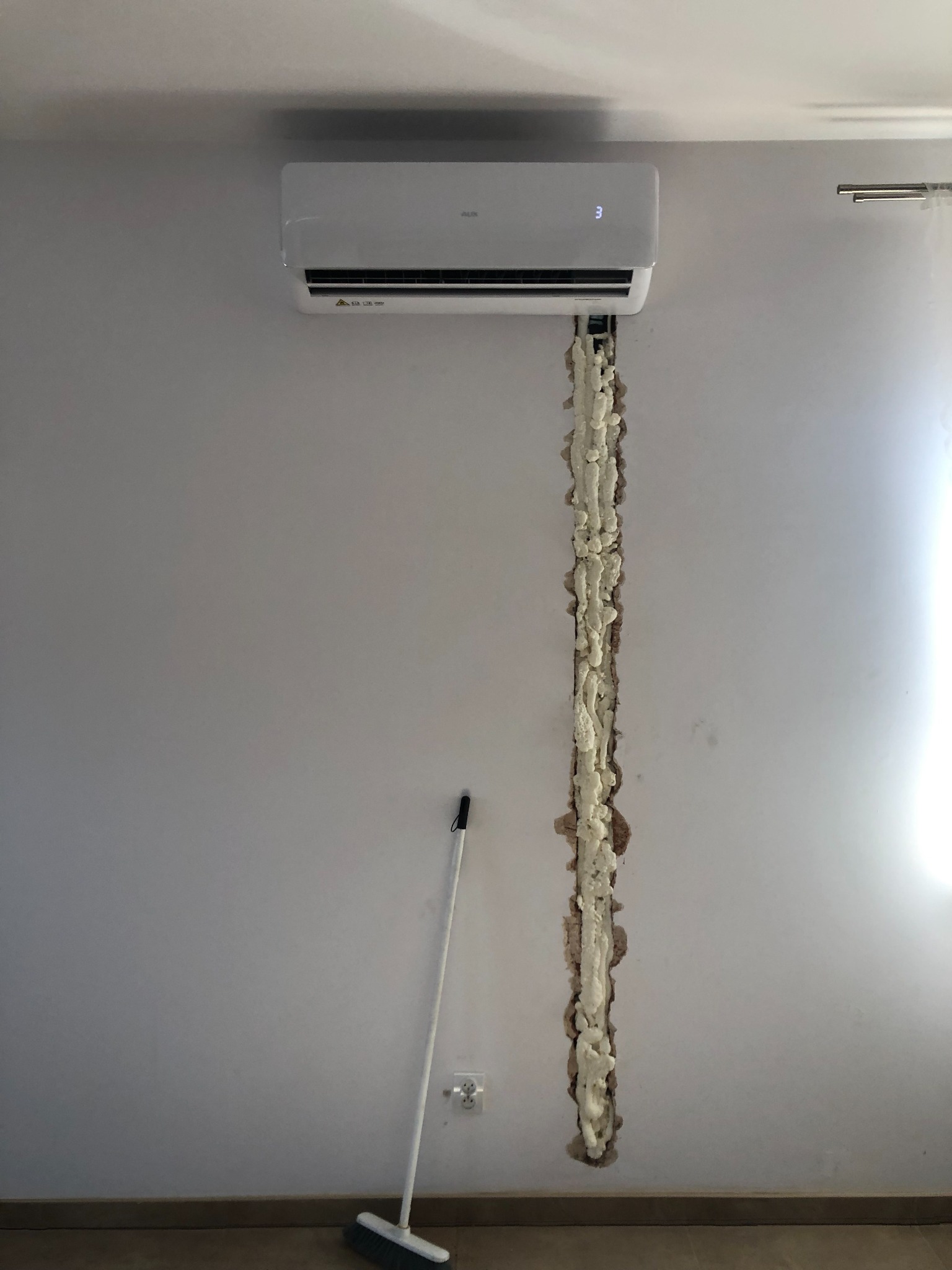 Montaż klimatyzatora ściennego w pomieszczeniu, widoczna biała jednostka wewnętrzna nad pionową bruzdą w ścianie wypełnioną pianką montażową; na podłodze stoi zmiotka.