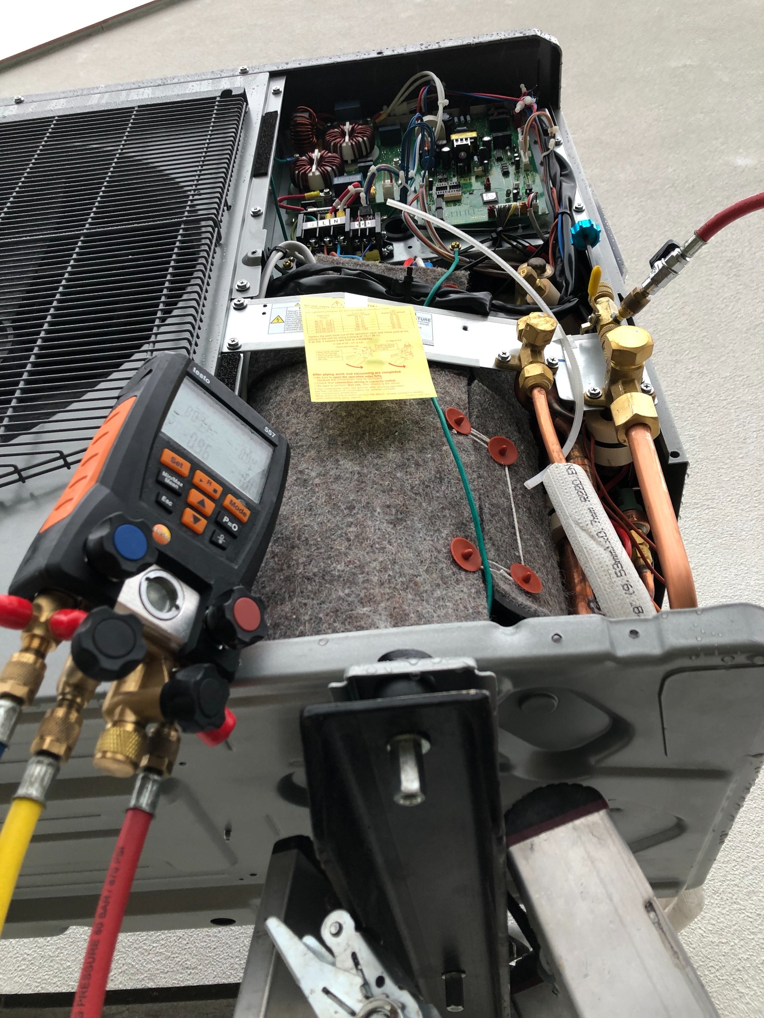 Otwarta obudowa zewnętrznej jednostki klimatyzacji podczas serwisu, widoczne podłączenie manometru cyfrowego Testo 557 i instalacja z rurami miedzianymi, żółta naklejka informacyjna i fragment...