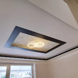 Nowoczesny sufit podwieszany z oświetleniem LED w kształcie trzech połączonych okręgów, otoczony czarną ramą i perforowaną płytą, widoczny w jasnym wnętrzu z oknem.