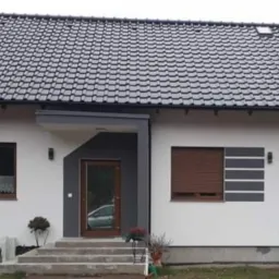 Nowoczesny dom jednorodzinny z białymi ścianami, ciemną dach&oacute;wką i szarymi elementami dekoracyjnymi przy oknach oraz brązowymi roletami zewnętrznymi, widok z zewnątrz.