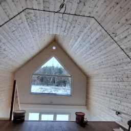 Poddasze wykończone boazerią z oknem trójkątnym, widok na zimowy krajobraz, widoczne krzesła i wiadro z farbą na drewnianej podłodze.