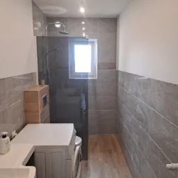 Nowoczesna łazienka z szarymi płytkami imitującymi marmur, kabiną prysznicową typu walk-in, drewnianą zabudową i pralką pod blatem.